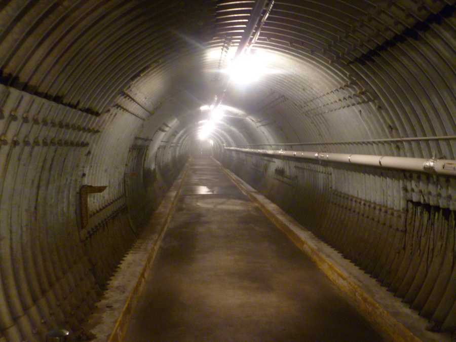 Empty corrugated-metal tunnel with ceiling lamps and an exposed pipe along one wall.