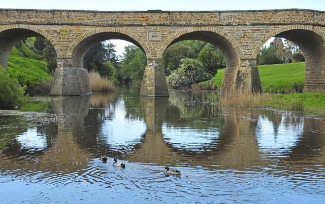 Several sandstone arches and piers cross a river, with ducks in the foreground and green banks beyond.