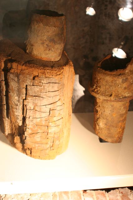 A cracked wooden pipe section stands beside shorter metal pipe sections in a glass-fronted display, with reflected lamps above.