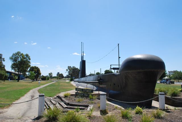 Black submarine casing and a tall fin stand amid paths and grass, with a small HMAS Otway sign near the rounded end.