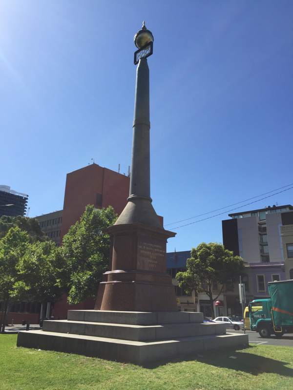 A tall granite column rises from a stepped base, with three eights and a globe at the top; buildings, trees and traffic stand behind it.
