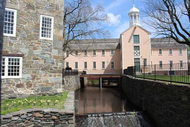 A stone-lined water channel passes beneath a footbridge toward a peach-colored mill with a bell tower; part of a stone mill stands at left.