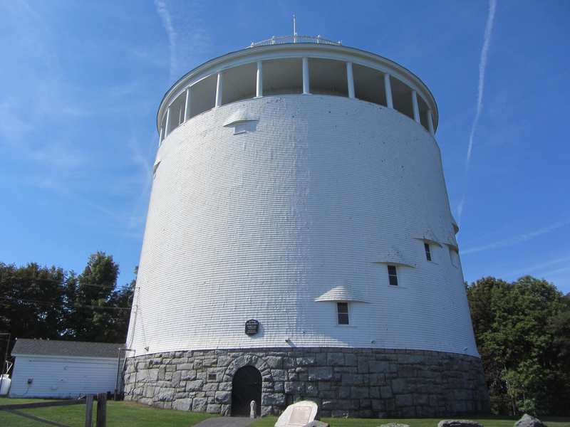 A white cylindrical shingled building on a stone base, with small hooded windows rising around its wall and a colonnaded upper deck.