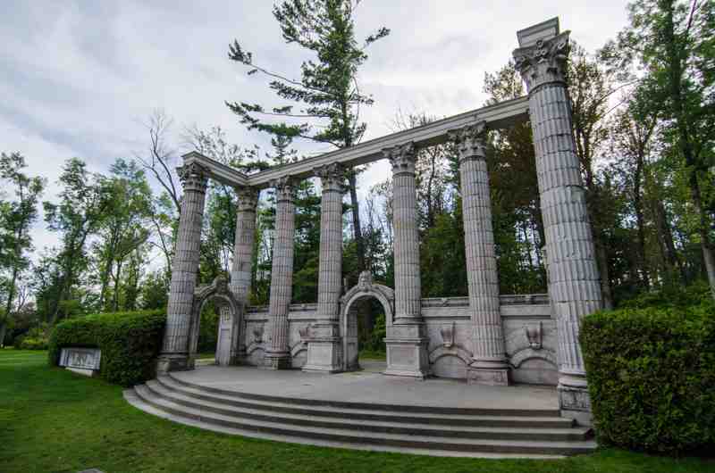 An empty curved stone stage and steps sit before tall fluted columns and three arches, with lawn and trees around them.