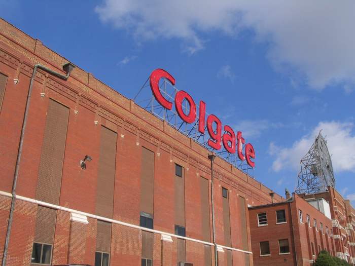 Red Colgate letters rise above a long brick industrial building; the rear steel framework of a large clock stands at the right.