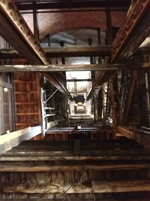 An upward view through successive beams and platforms inside a brick tower, with daylight visible higher up.