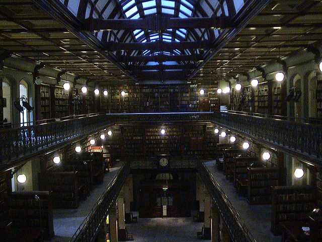 Two levels of galleries border a long library chamber beneath a glazed roof; globe lights glow along the balconies.