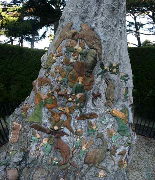 Painted fairies, birds and animals cover a broad face of a pale tree trunk, with a garden fence and foliage behind it.