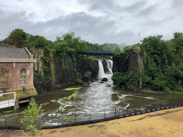 A brick powerhouse stands at the left of a rocky, tree-lined gorge, with a waterfall beneath an arched metal bridge.