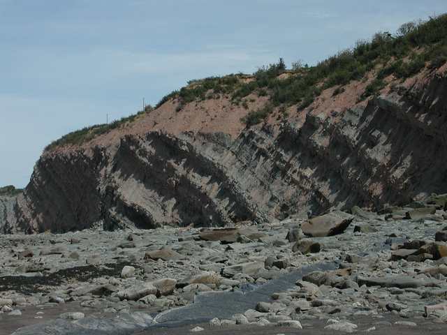 Tilted rock layers form a coastal cliff above a rocky beach, with vegetation along the top.