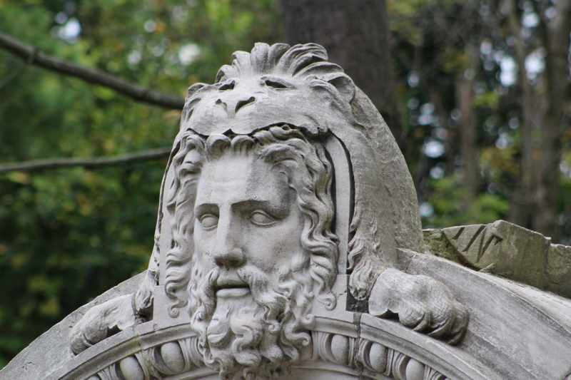 A close view shows a bearded stone face beneath a carved lion’s head above an arch, with green trees behind.