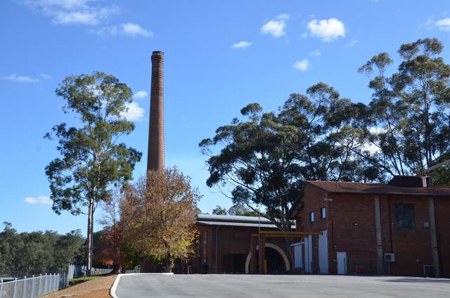 A tall brick chimney above a low red-brick pumping-station building, with an arched opening and trees under a blue sky.