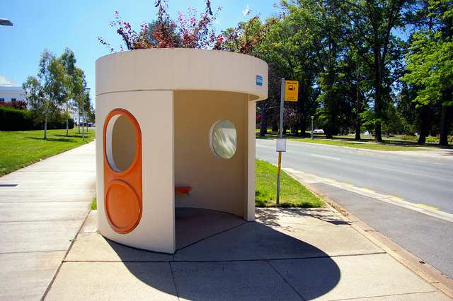 Cream concrete cylindrical bus shelter with orange window surrounds, entrance and nearby bus-stop sign.