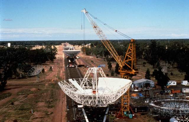 A crane beside a partly assembled radio dish, with parallel rails and more dishes extending into the distance.