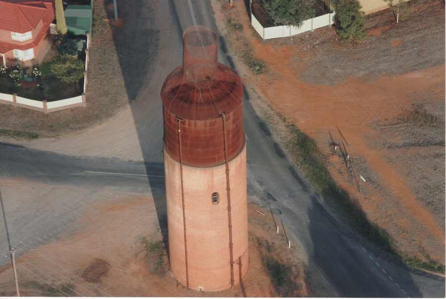 An aerial view shows a tall brick water tower with a red tank and open mesh shoulders and neck, surrounded by streets and houses.