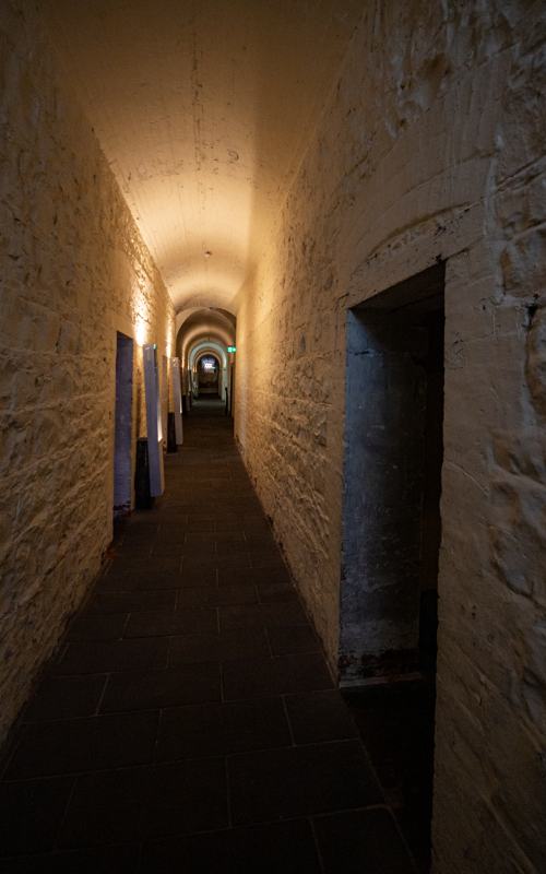 A narrow passage has pale rough walls, an arched ceiling, dark paving and open doorways, with warm lights and modern exit signs.
