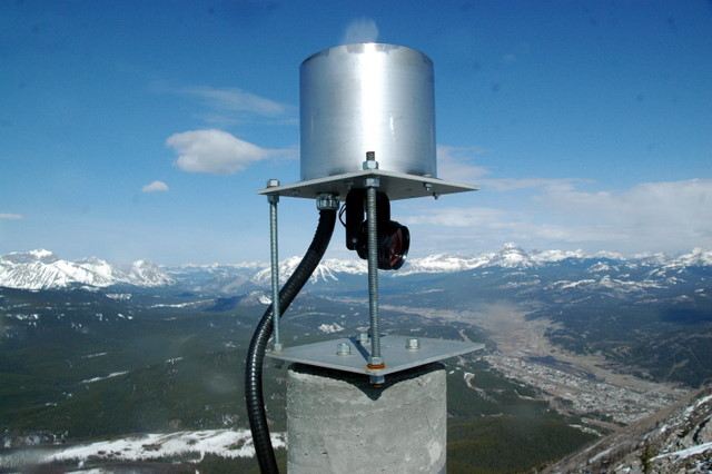 A small reflector and cables sit beneath a metal cylinder on a concrete pillar above a snowy valley.