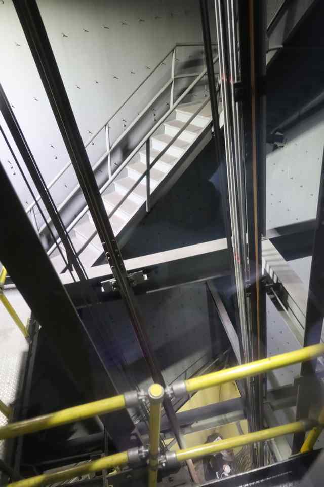 A view through the front of a tram toward interior steelwork, stairs, rails and yellow railings inside the Arch.
