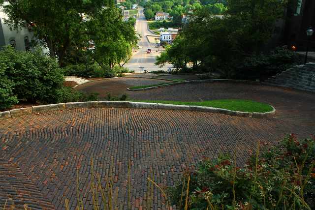 A brick street winding downhill around planted islands edged with stone curbs, with buildings and another street beyond.