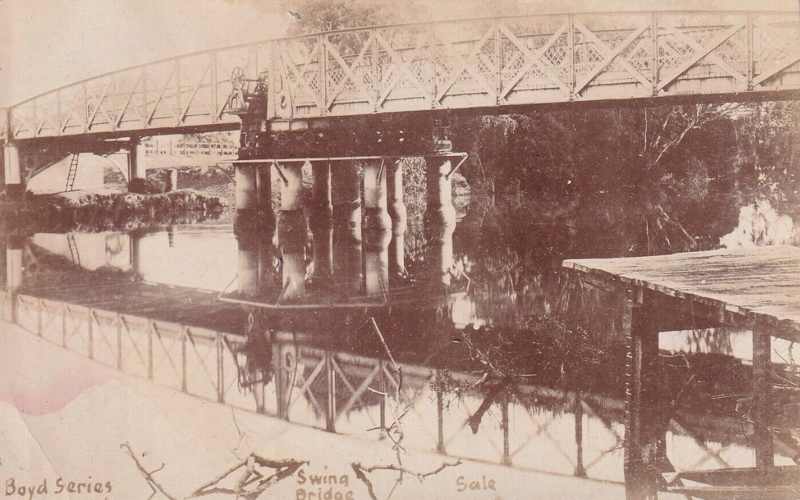 Sepia postcard of the iron bridge across the river, with its central supports and lattice railings reflected in the water; the original postcard lettering remains visible.