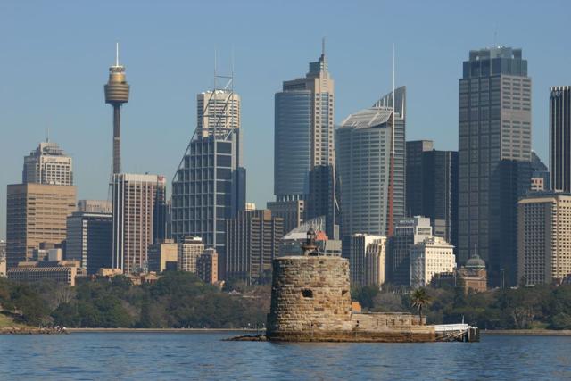 A low stone fort and round tower stand in blue harbour water, with Sydney’s tall buildings behind.
