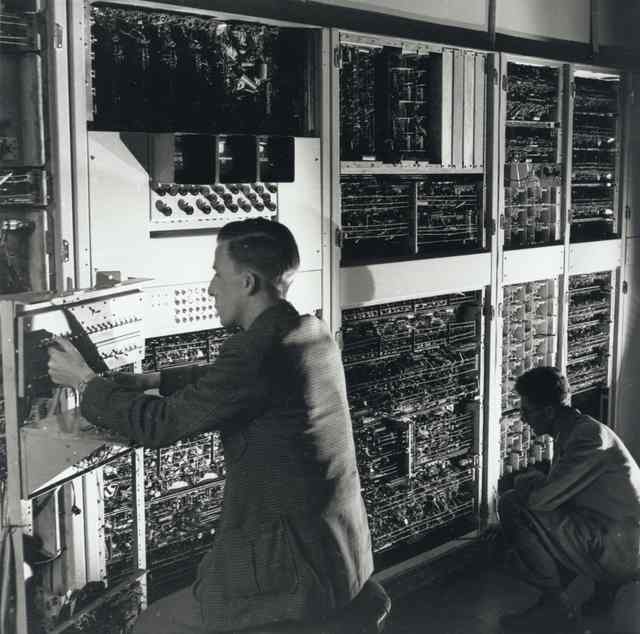 Two people work beside exposed computer racks and controls in a closer oblique archival view.