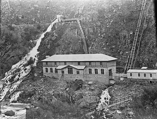A grainy black-and-white photograph shows a long powerhouse beneath a rocky slope, with a large pipe descending behind it, steep steps at the right and a suspension bridge below.