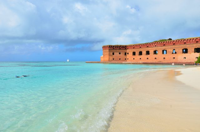 Fort Jefferson’s long brick wall stands beside clear shallow water and a sandy beach.