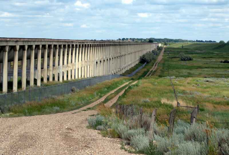 A long row of concrete aqueduct supports recedes across a grassy valley beside a fence and dirt track.