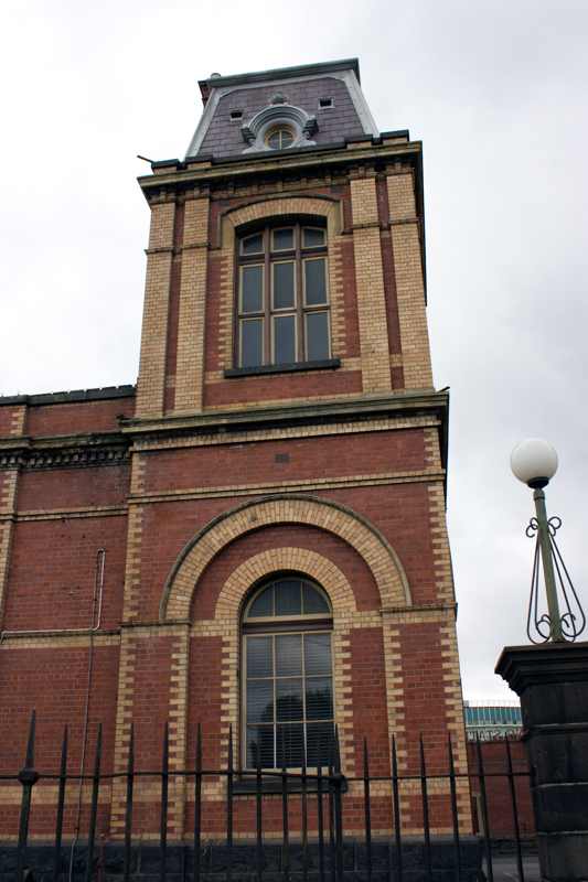 A red-and-cream brick tower rises above arched windows, with a steep dark roof, round upper window and iron fence in front.