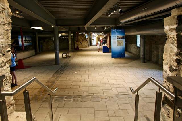 Steps lead into a broad underground room with stone walls, metal columns, overhead beams and small display panels.