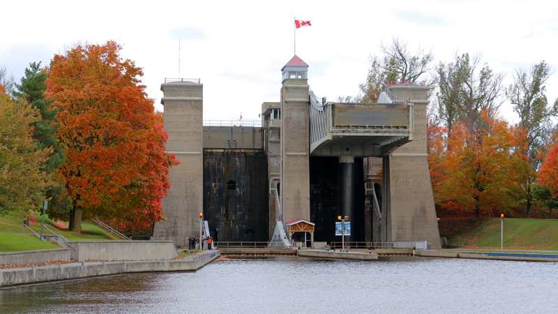 A raised metal boat chamber rests on a dark cylindrical ram between concrete towers, with autumn trees and the canal in front.