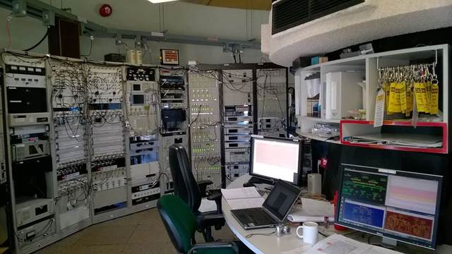 An empty control room contains equipment racks with many cables, computer screens, a laptop, chairs and a mug.