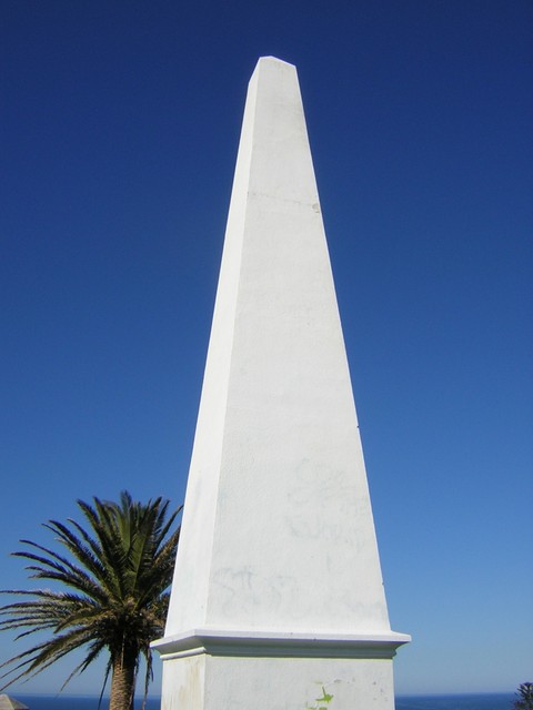 A pale tapered obelisk rises against blue sky, with a palm at left and a narrow strip of sea behind its lower portion.