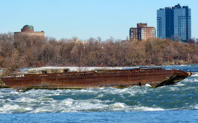A long rusted scow lies across churning river water, with trees and buildings beyond.