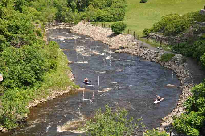 Two paddlers in a narrow rocky channel beneath suspended slalom gates, with trees and a fenced path along the banks.