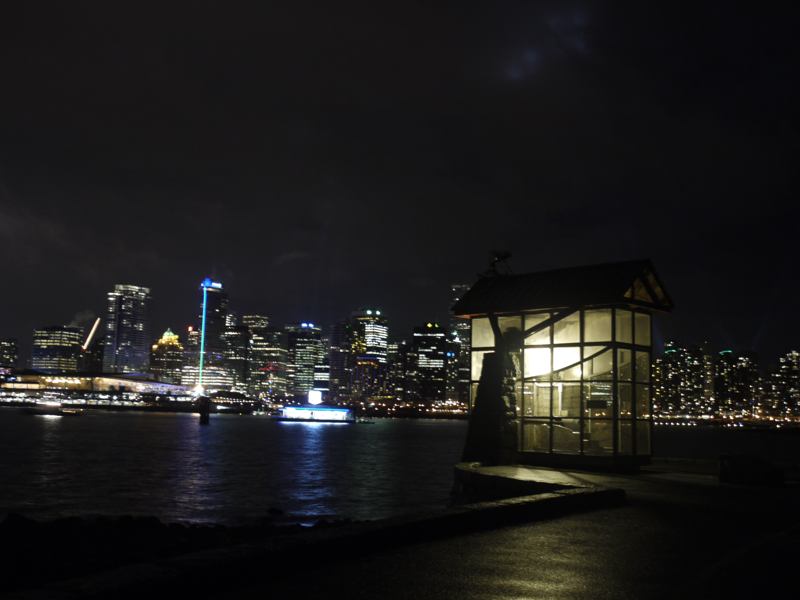 An illuminated gun enclosure stands beside dark water, with Vancouver’s lit buildings beyond.
