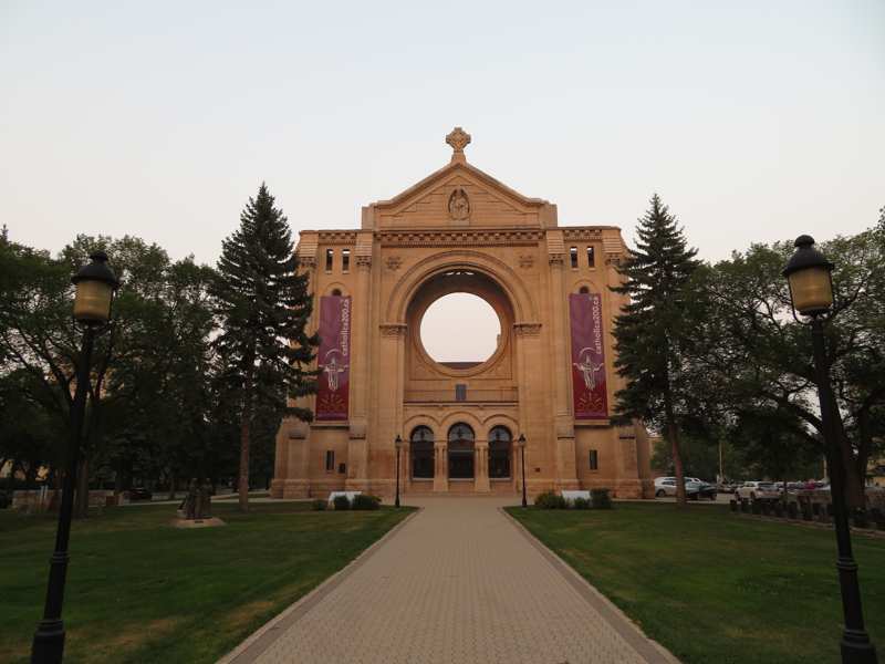 A straight path leads toward the old cathedral’s stone façade, with three arched entrances and a large empty circular window.