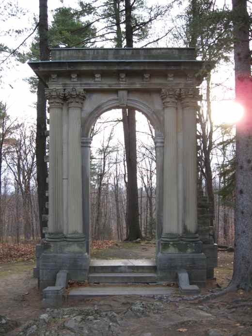 A freestanding stone arch with four carved columns and steps, surrounded by woodland.