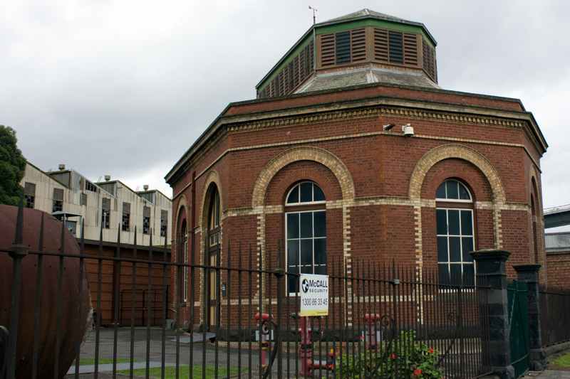 A many-sided red-brick building has cream-brick window arches and a raised louvred roof, behind a black fence.