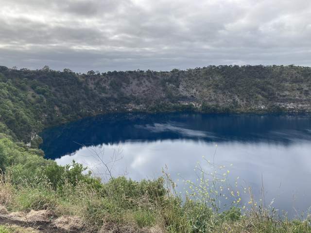 A crater lake reflecting grey clouds, with green shrubs in the foreground and a steep tree-covered opposite shore.