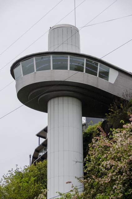 Close upward view of the rounded observation deck, its angled windows and cylindrical shaft, with vegetation and overhead wires.