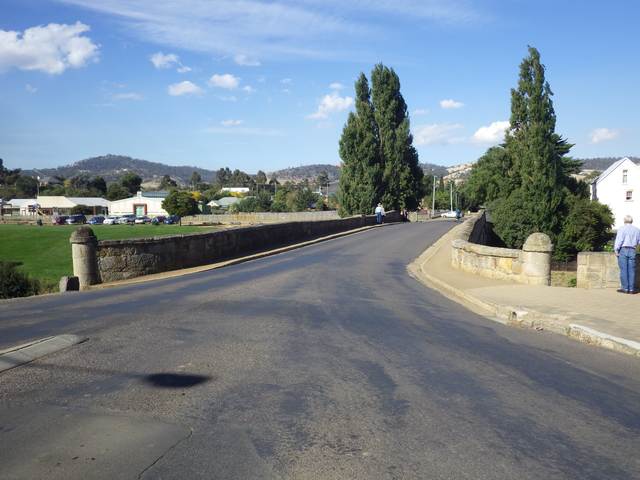 A road crosses between stone parapets with narrow footpaths; a few pedestrians appear along the sides.