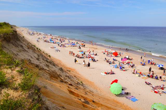 A long sandy beach with small groups of people lies between a steep bluff and the sea.