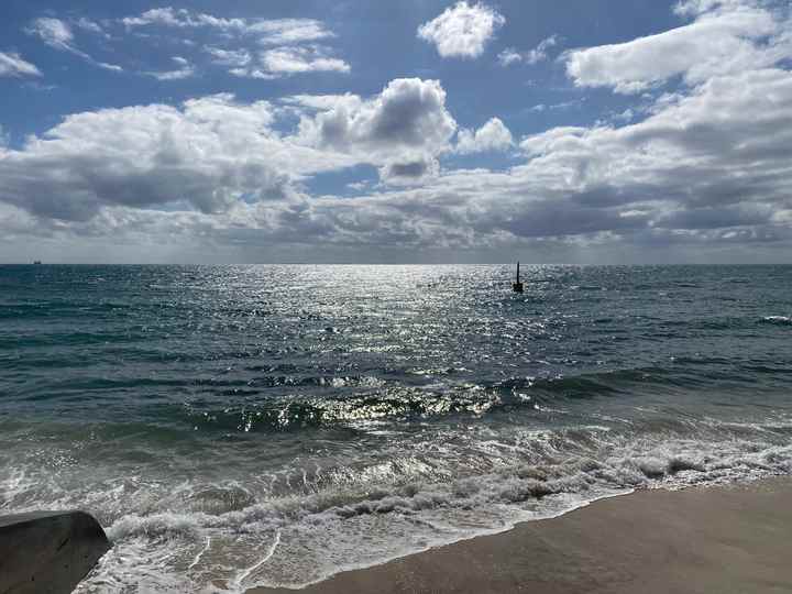 The upright pylon stands offshore beyond sunlit water and breaking waves on Cottesloe Beach.