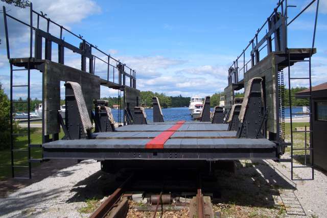 An empty wooden-decked railway carriage has tall sides, padded uprights and visible wheels on rails, with boats and water behind.