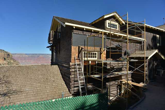 Metal scaffolding crosses a brown building with pale window trim, exposed wall areas and lighter shingles, with the canyon behind it.