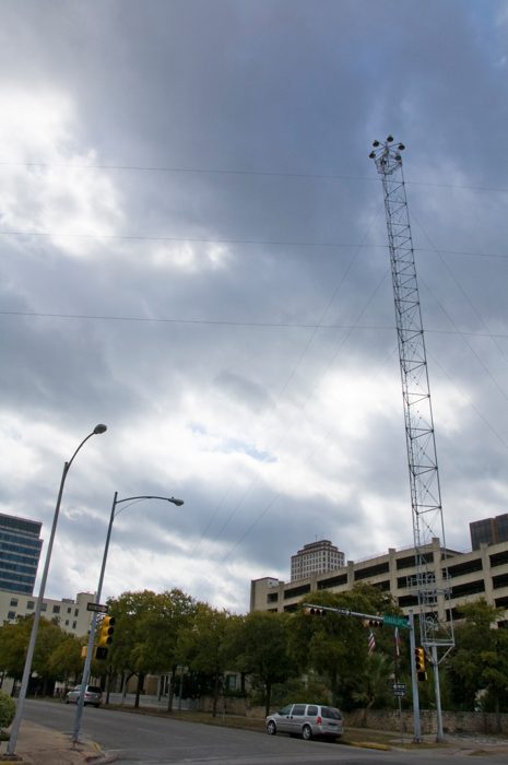 A tall lattice lighting tower beside a downtown street, with lamps above nearby buildings.