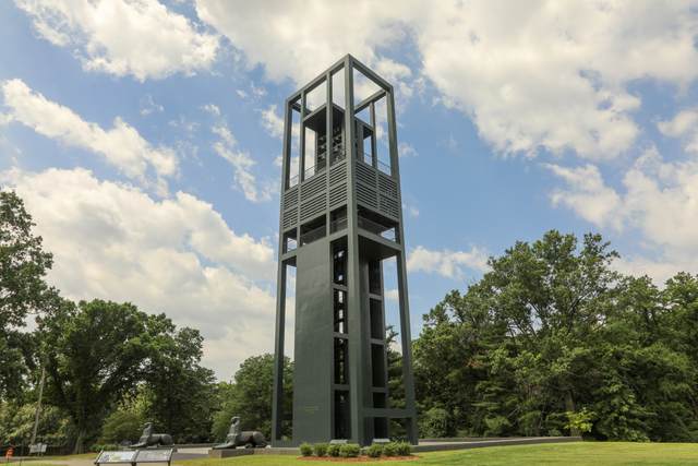 A rectangular open steel bell tower rises over grass and trees, with two low lion sculptures beside its base.