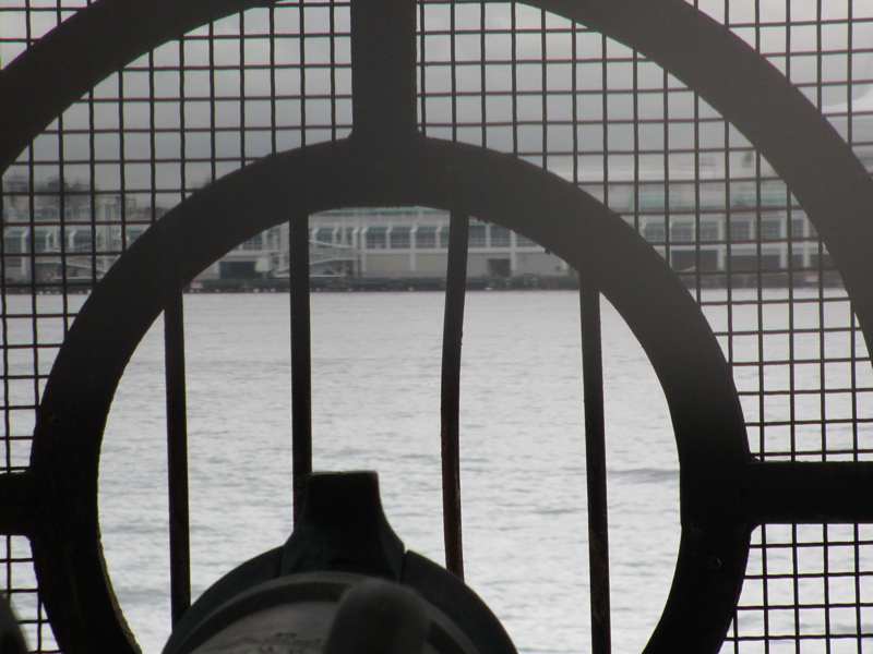 The end of a gun barrel appears below curved bars and square mesh, with water and a building beyond.